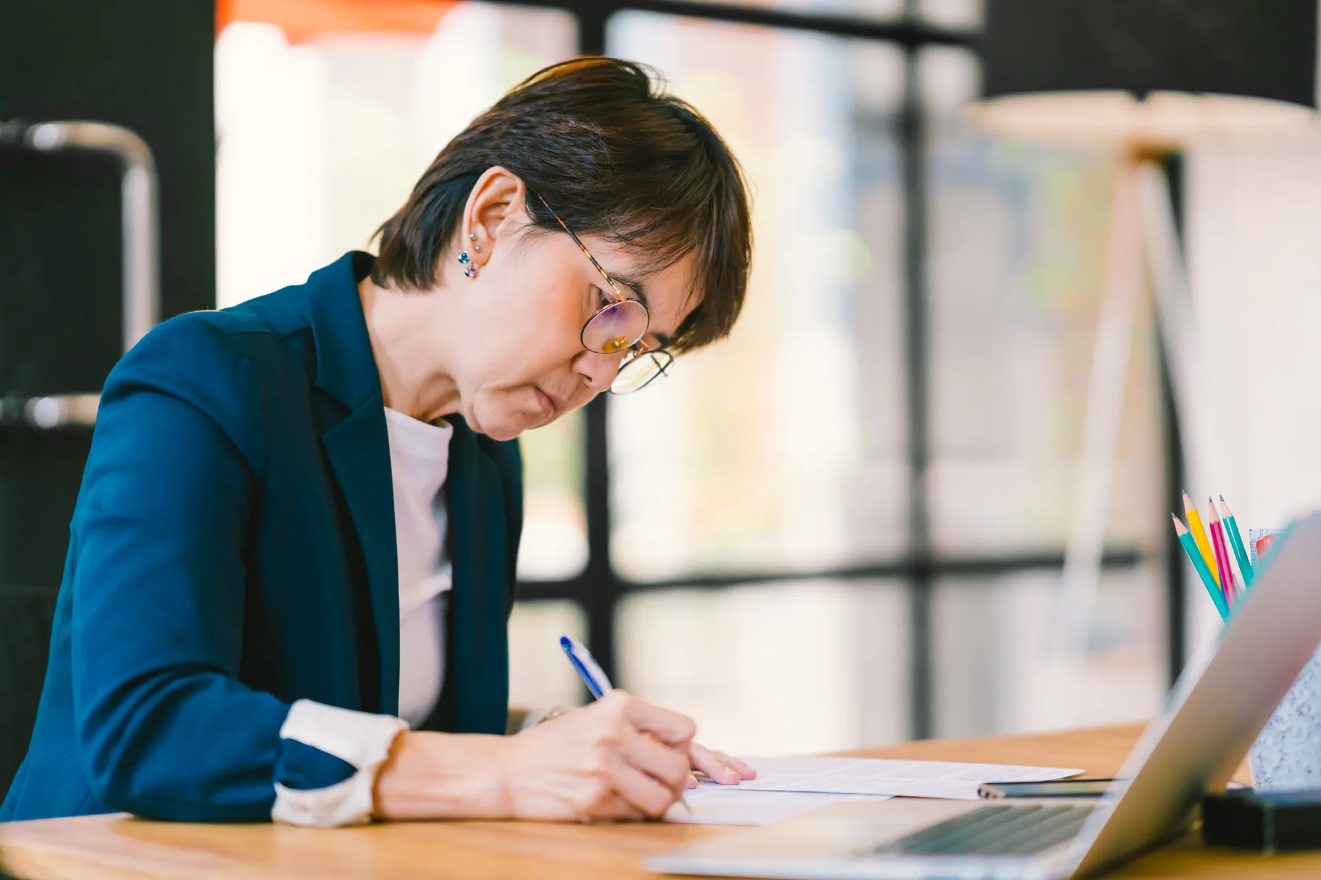 A professional woman in glasses sits at a laptop, writing on a sheet of paper.