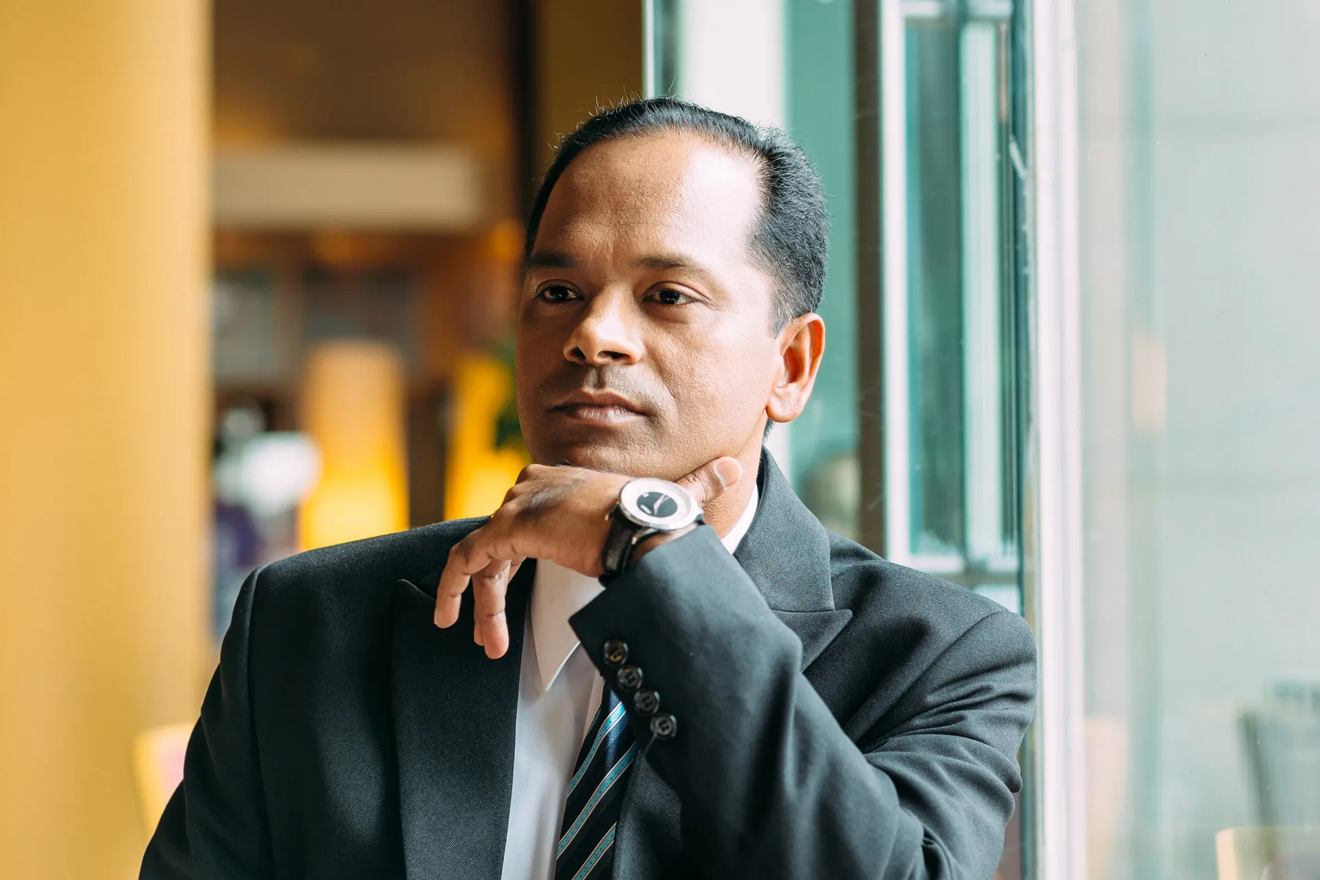 A professional man in a suit and tie sitting by a window, engrossed in his thoughts.