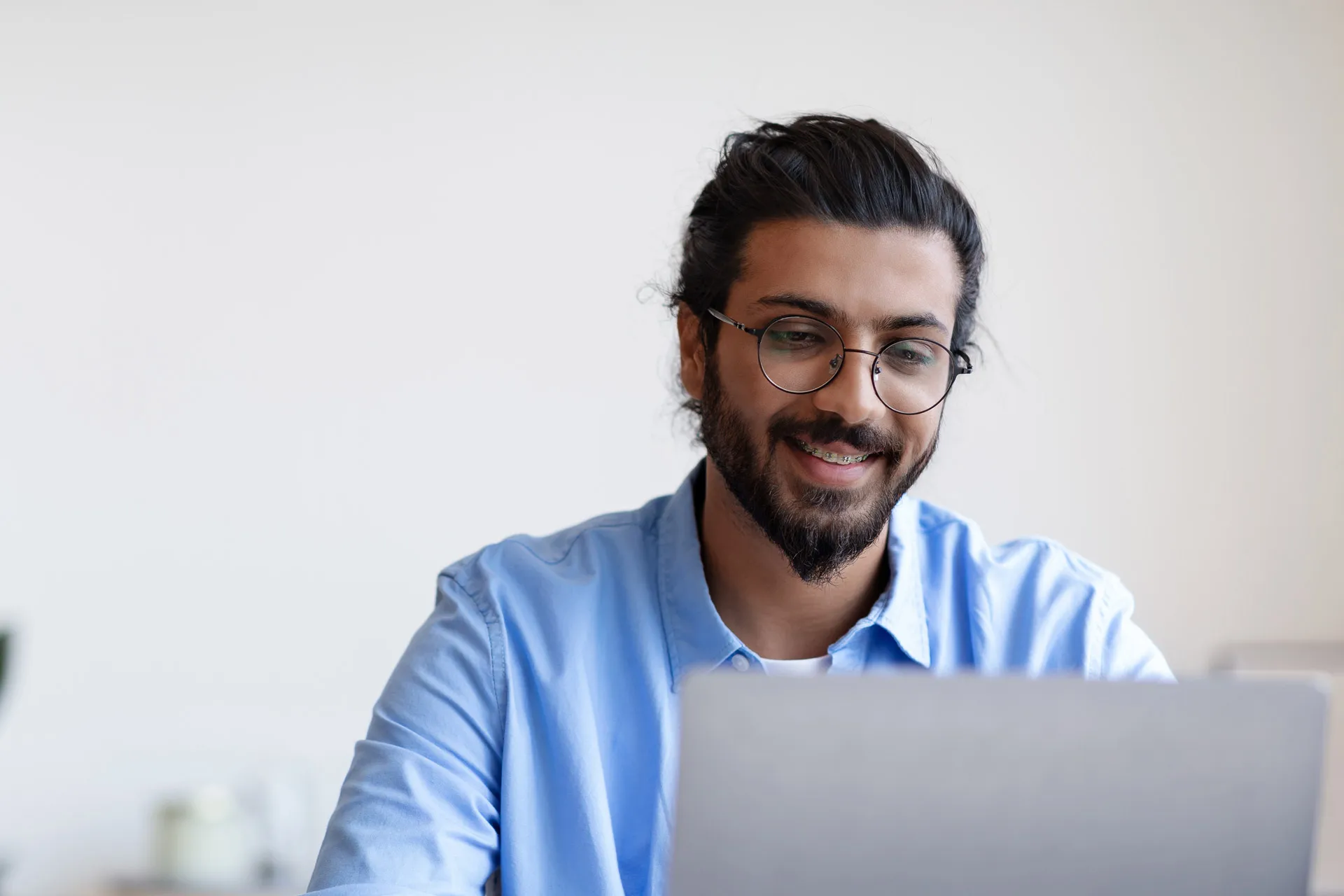 A bearded young man wearing glasses smiles while working on a laptop.