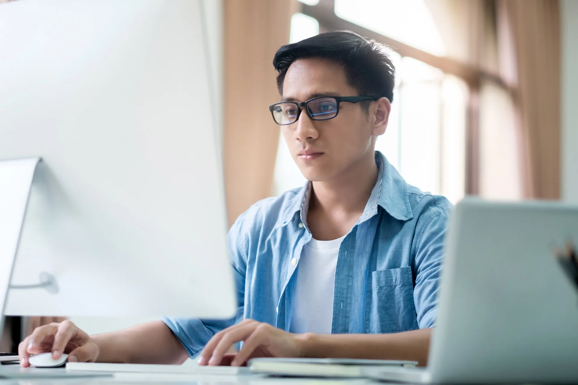 An Asian man focused on his work, sat at a computer in his office.