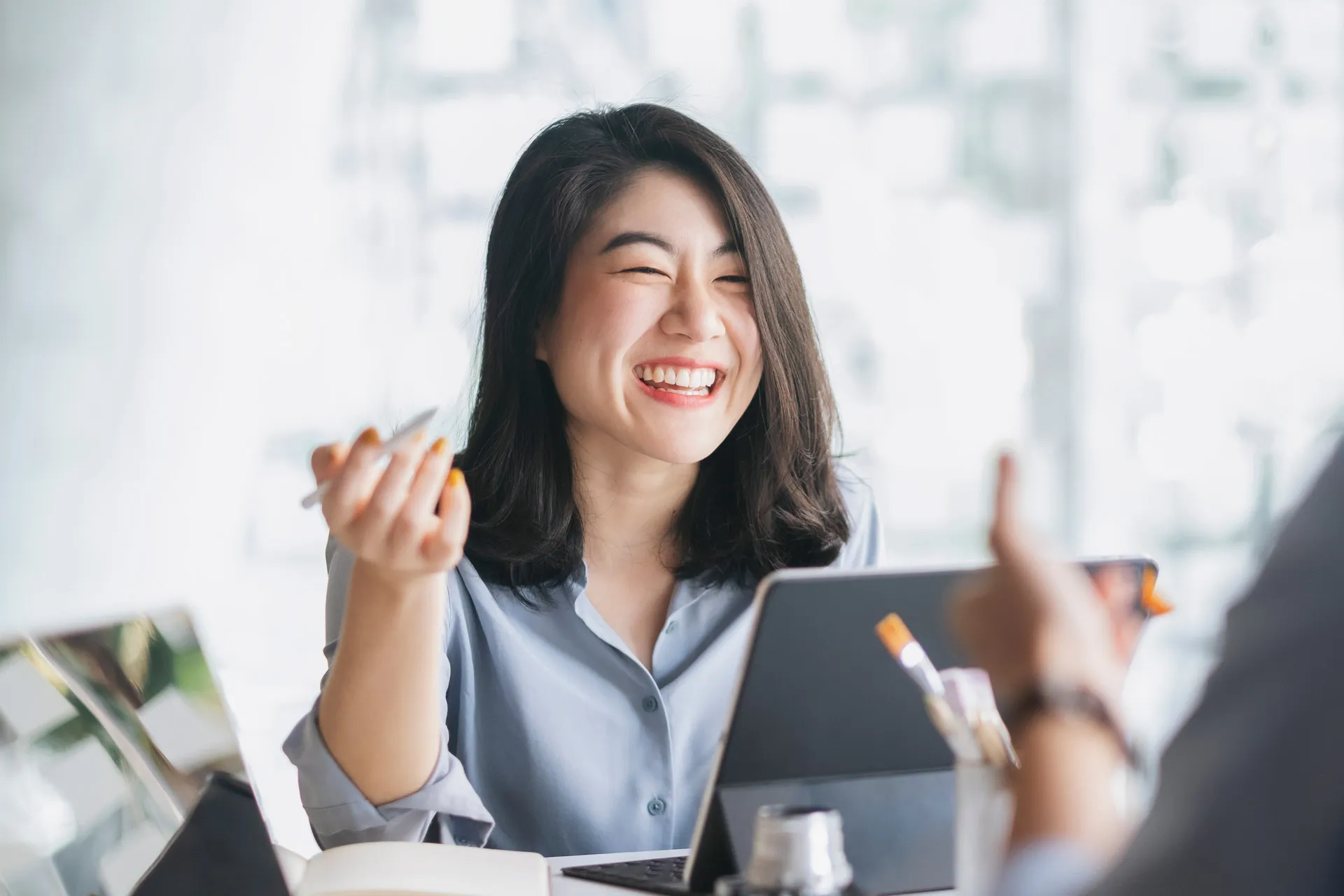 A smiling Asian woman works on her laptop, while engaging in conversation with a colleague.