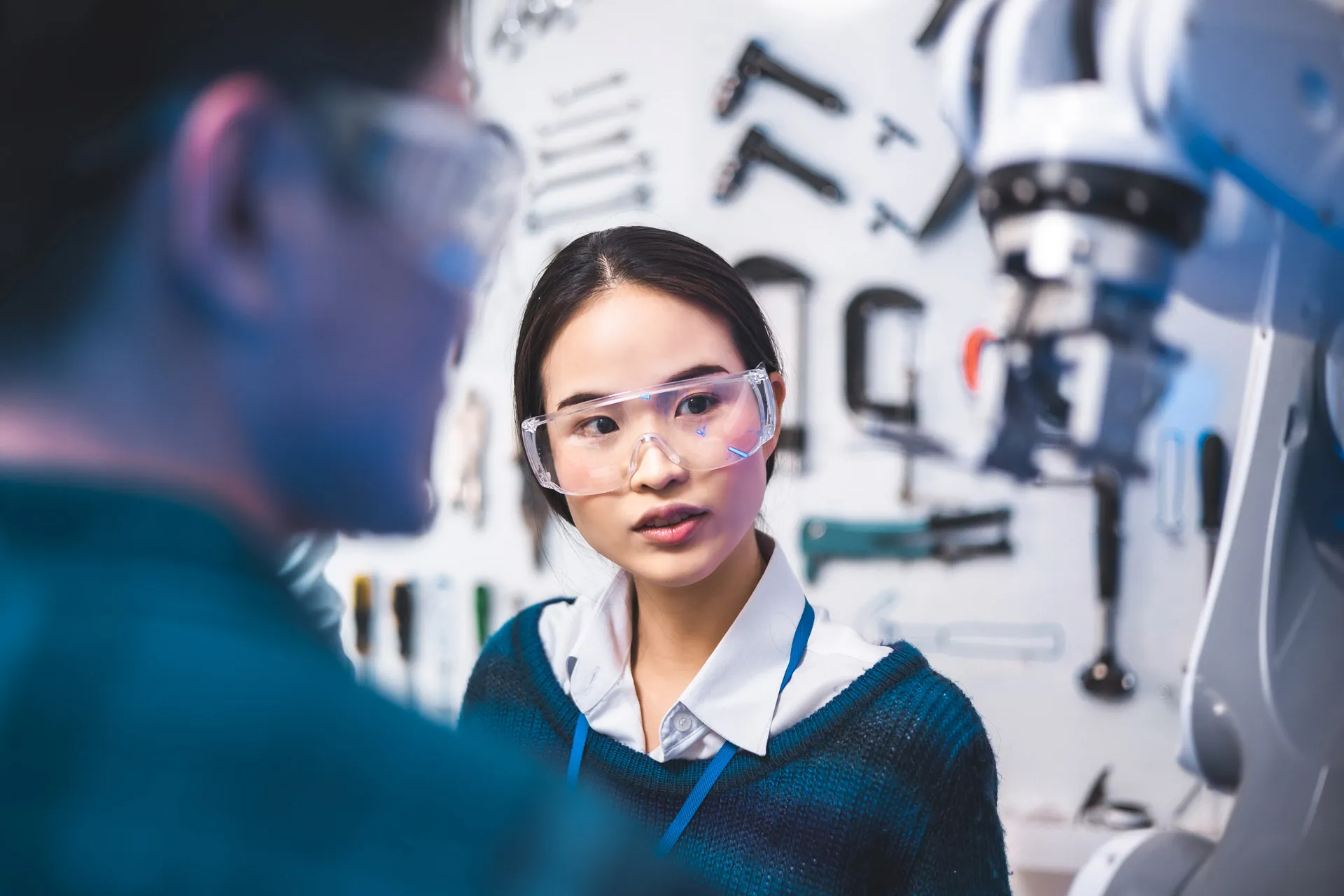 A woman wearing safety glasses examines a robot in a technology workshop.