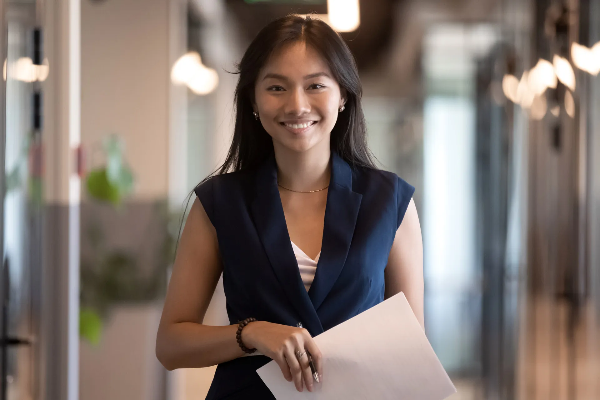 Smiling young woman in business attire holds a paper and pen in a contemporary office setting.