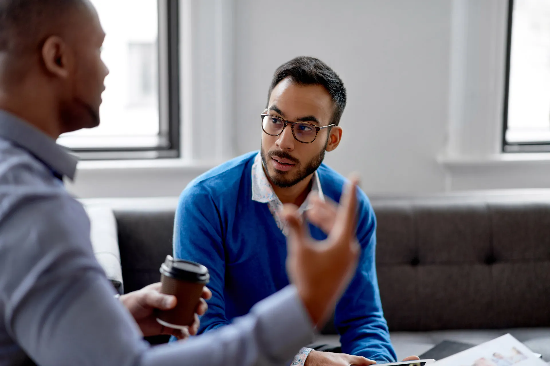 Two men drinking coffee, engaged in a conversation in a professional office environment.